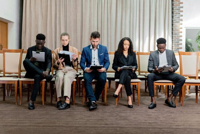 Five people wearing business attire, sitting on a row of chairs in a straight line, looking down at clipboards.