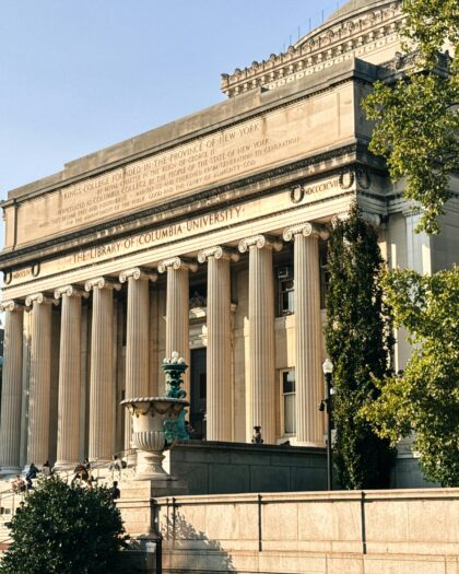 The image shows the grand entrance of a library of columbia university building with tall columns and an inscription. People are sitting on the sunlit steps, surrounded by trees.