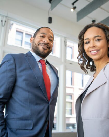Man and woman in business attire looking direct to camera with modern windows in the background