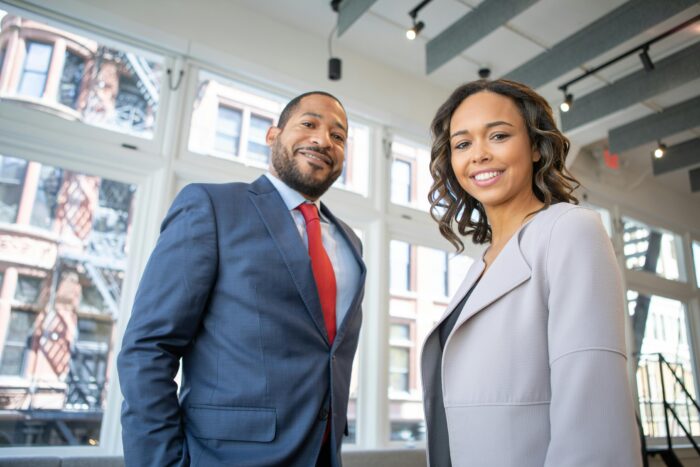 Man and woman in business attire looking direct to camera with modern windows in the background