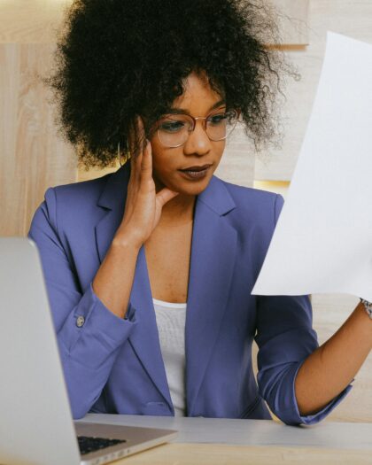 Woman sitting at desk with laptop, looking at paper in hand