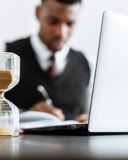 Picture of hour glass and laptop on a desk with man in suit blurred in the background
