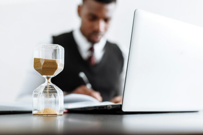 Picture of hour glass and laptop on a desk with man in suit blurred in the background