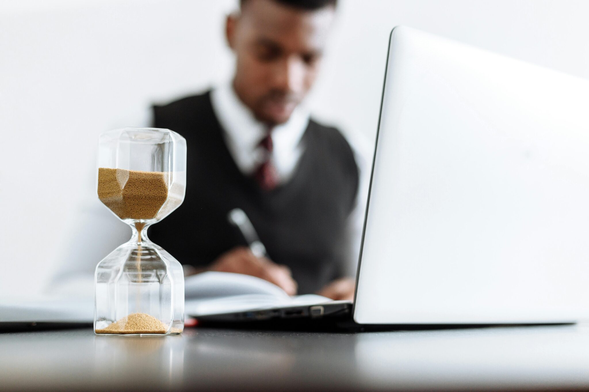 Picture of hour glass and laptop on a desk with man in suit blurred in the background