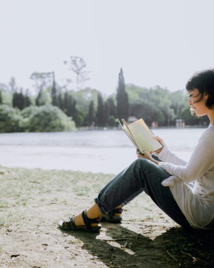 A young girl sitting under a tree by a river, reading a book peacefully