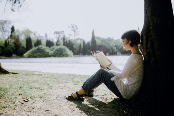 A young girl sitting under a tree by a river, reading a book peacefully
