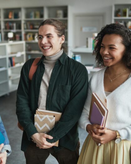 Two young girls and one young boy standing in a library, holding books and laughing together while discussion.