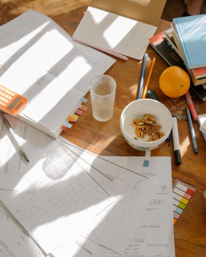 A round wooden table with papers and books strewn about, with a few mugs.