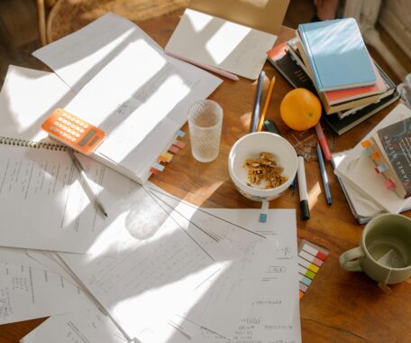 A round wooden table with papers and books strewn about, with a few mugs.