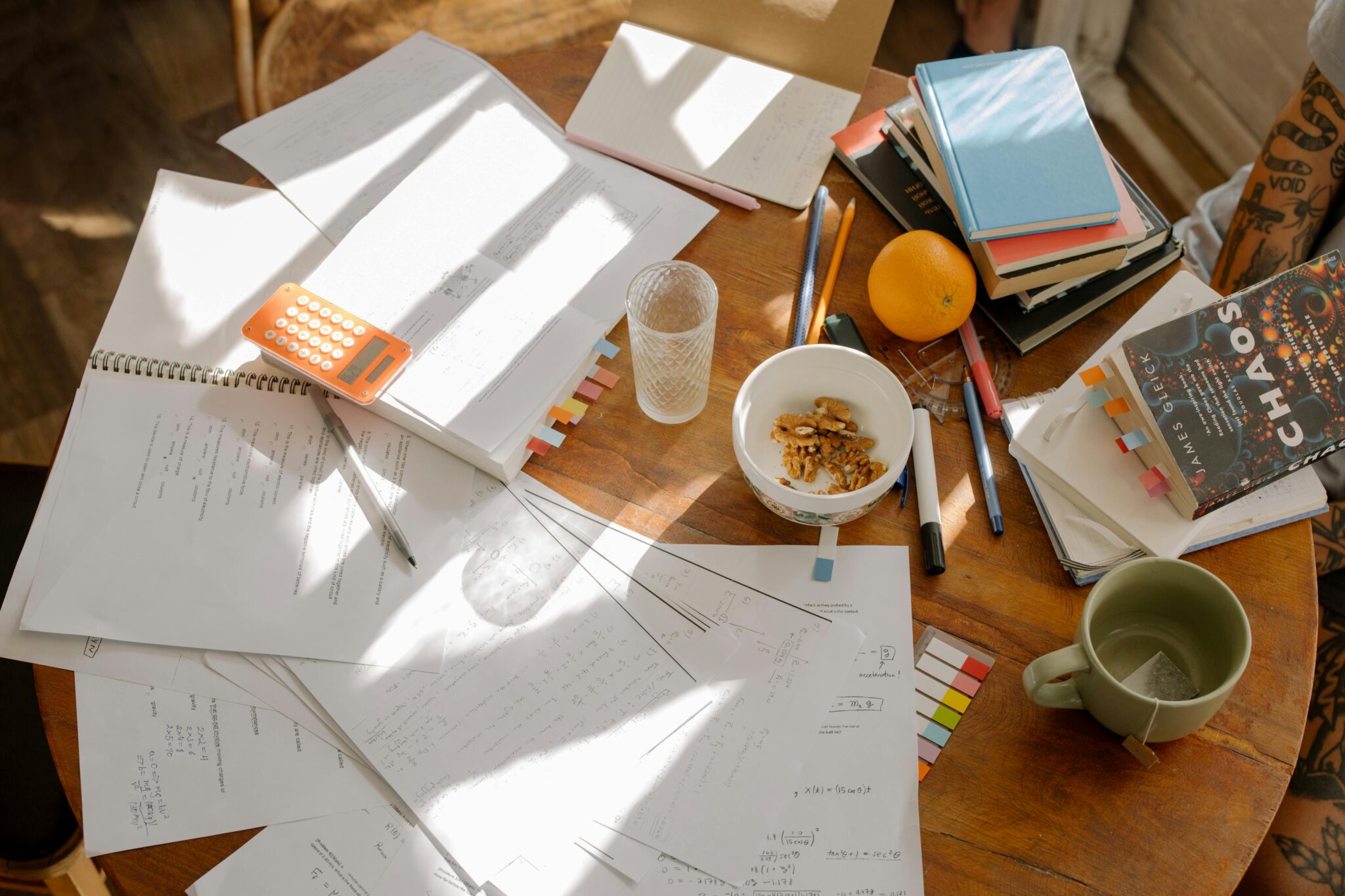 A round wooden table with papers and books strewn about, with a few mugs.