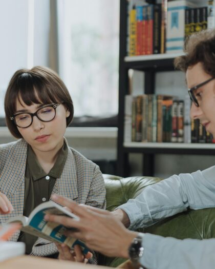 A young boy and girl sitting on a sofa, with the boy teaching the girl from an open book in his hand.