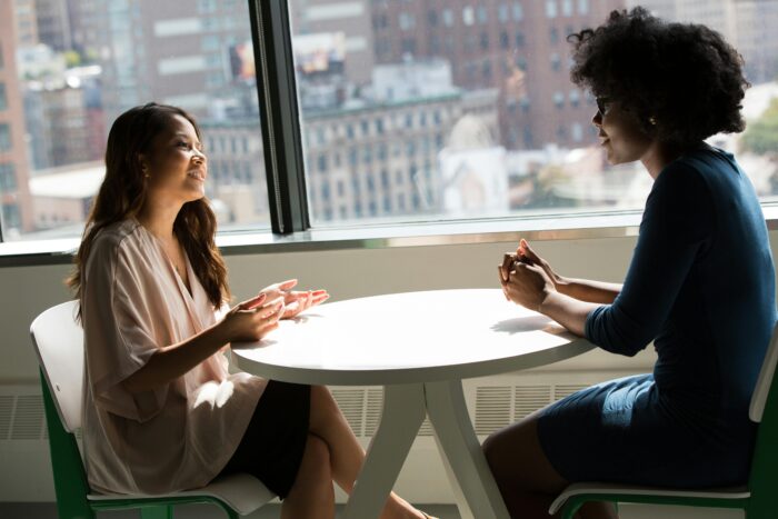 Two women in business attire discussing at a table next to a window