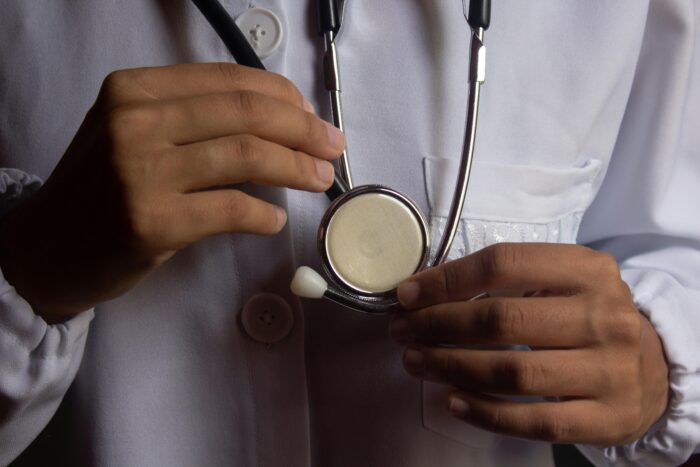A young male doctor in white scrubs stands facing the camera, with a stethoscope visible around his neck