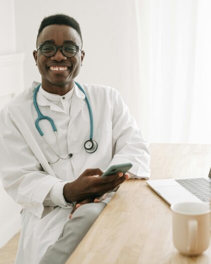 A young male doctor wearing a stethoscope around his neck smiles while looking at someone and holding a smartphone in his hand.