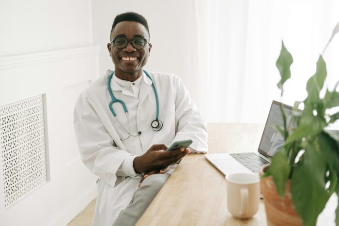 A young male doctor wearing a stethoscope around his neck smiles while looking at someone and holding a smartphone in his hand.
