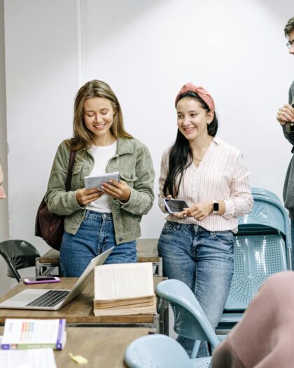 A group of five young adults in casual attire gather around a desk with open books and a laptop, smiling and chatting in a classroom setting.