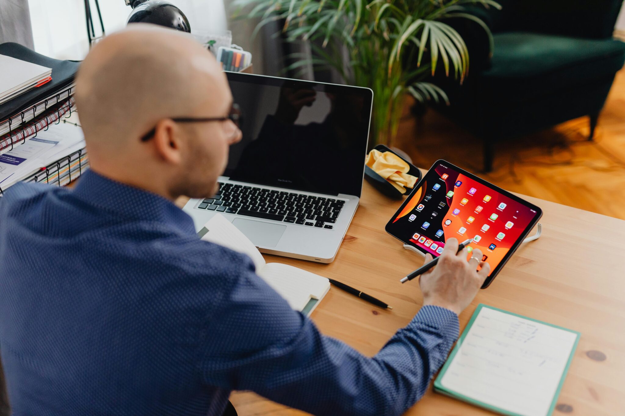 A man in a blue shirt uses a stylus on a tablet at a wooden desk. A laptop, open notebook, and papers are nearby. The setting is an organized home office.