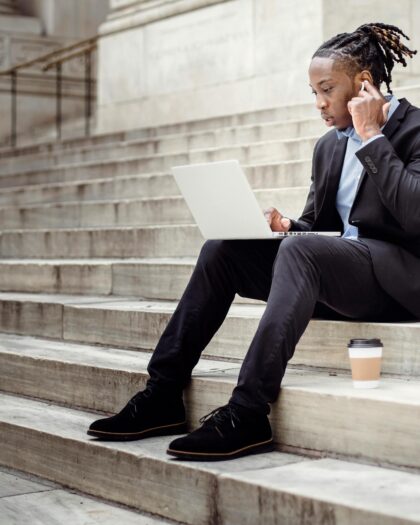Young man in business attire sitting on stone steps outside with laptop on lap