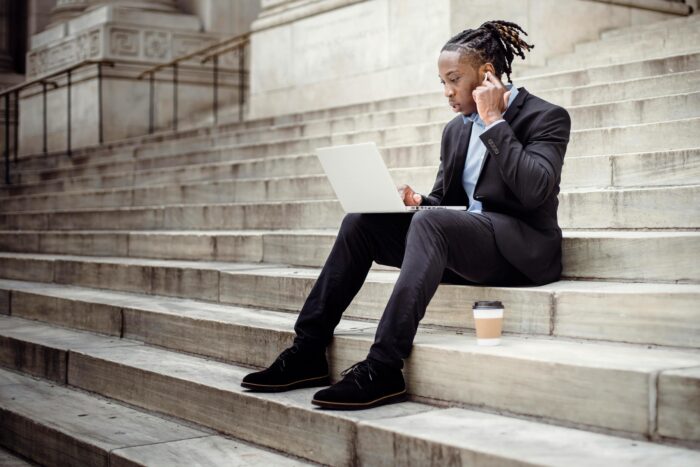 Young man in business attire sitting on stone steps outside with laptop on lap