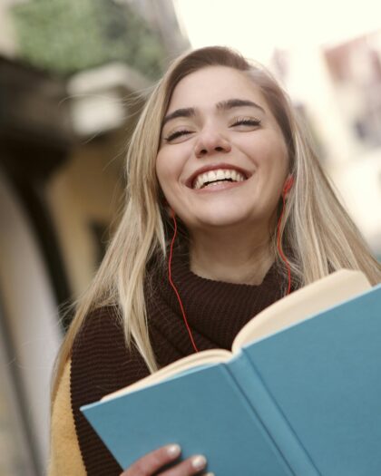 A young woman standing outside a university, smiling and reading a book.