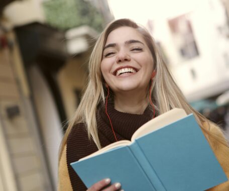 A young woman standing outside a university, smiling and reading a book.