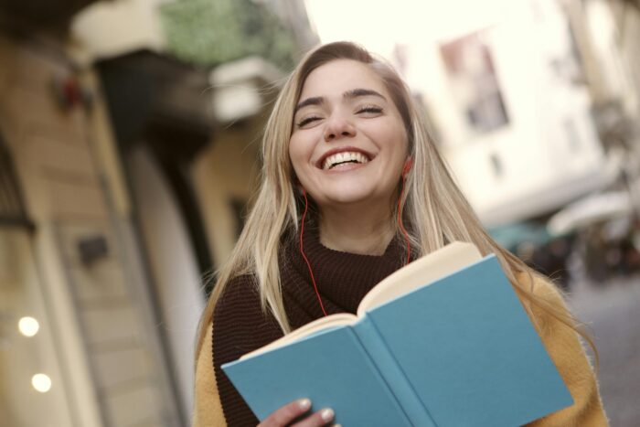 A young woman standing outside a university, smiling and reading a book.