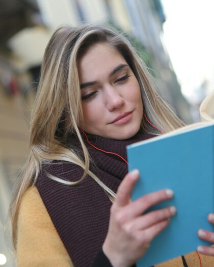 A young woman standing outside a university, smiling and reading a book.