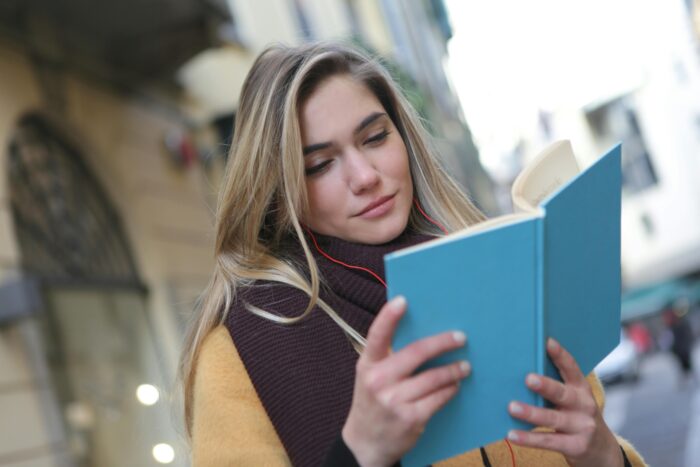A young woman standing outside a university, smiling and reading a book.
