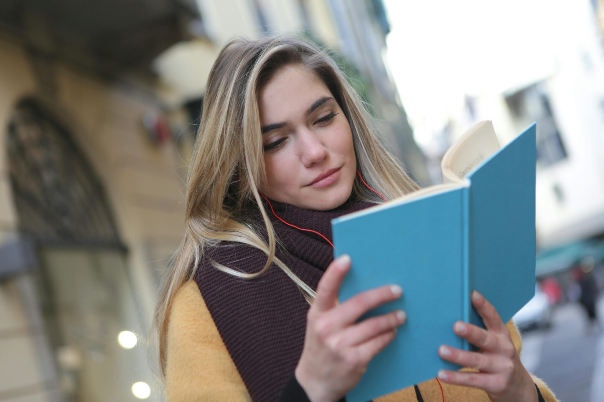 A young woman standing outside a university, smiling and reading a book.