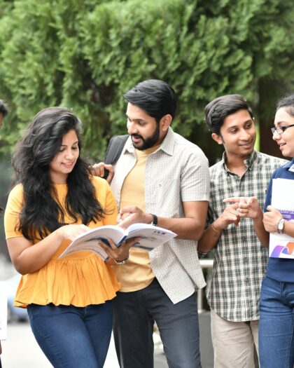 A group of five young adults standing outdoors, smiling and engaging with each other. One holds an open book; others carry folders. Lush greenery in the background.