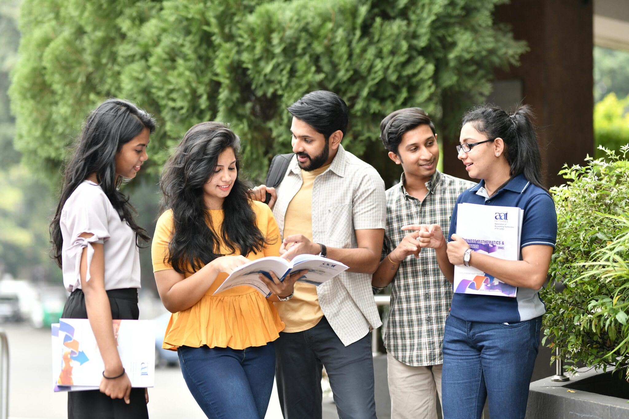 A group of five young adults standing outdoors, smiling and engaging with each other. One holds an open book; others carry folders. Lush greenery in the background.