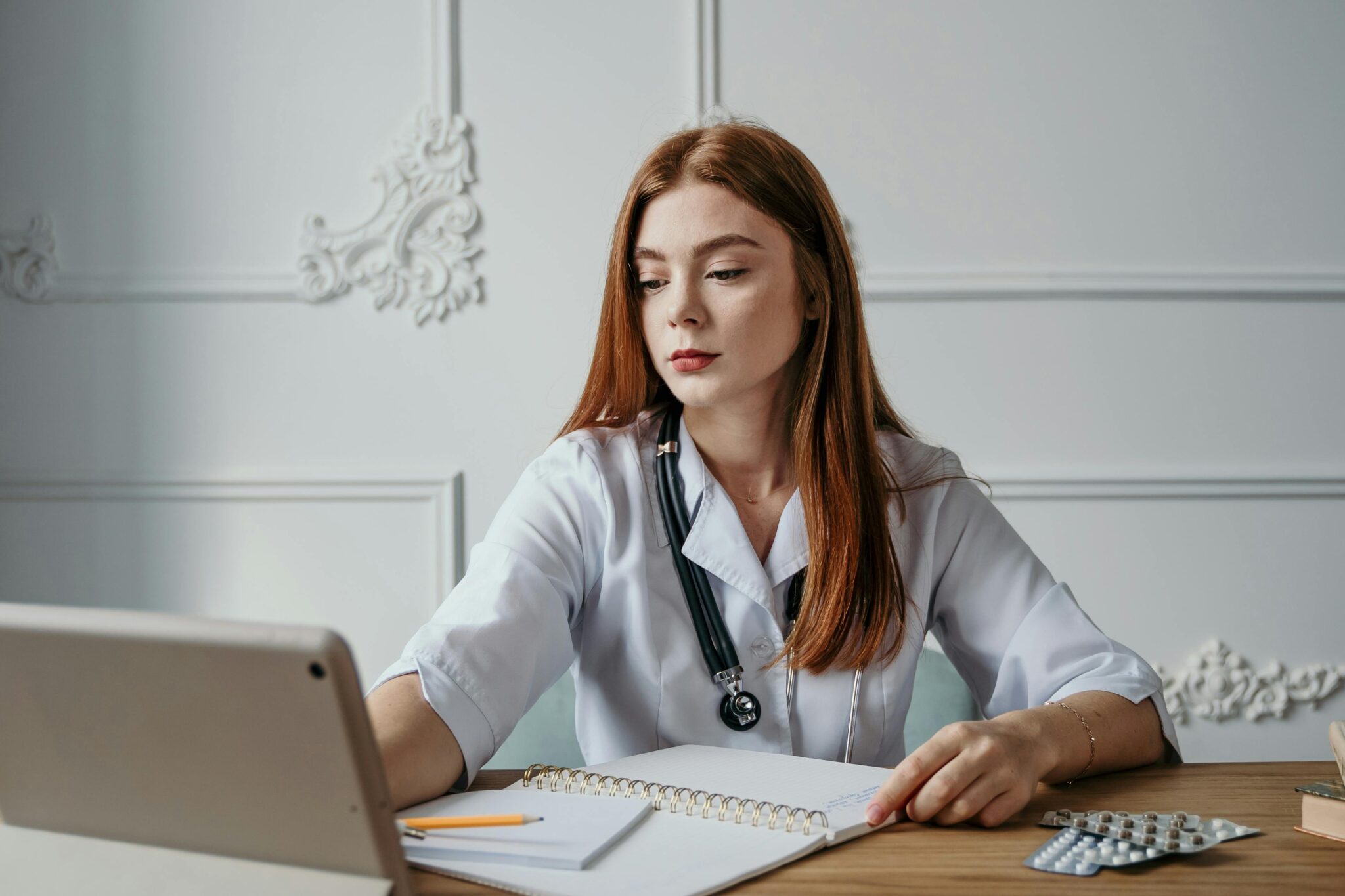 A young medical student studying about medicine on a tablet while taking notes in a notebook, with medicine on the table.
