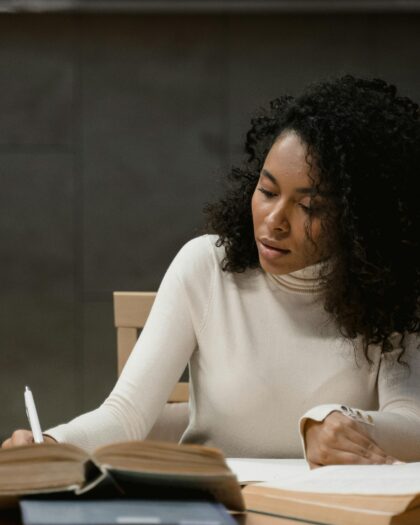 A young woman sitting at a study table, writing in a notebook surrounded by open books, focused on her study.