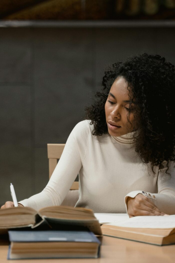 A young woman sitting at a study table, writing in a notebook surrounded by open books, focused on her study.