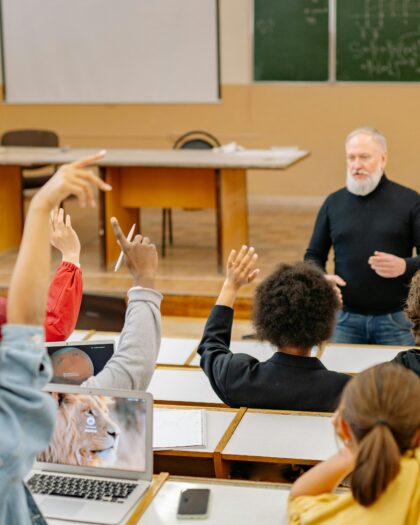 A photo of classroom where the teacher is asking a question, and the students are raising their hands to respond.