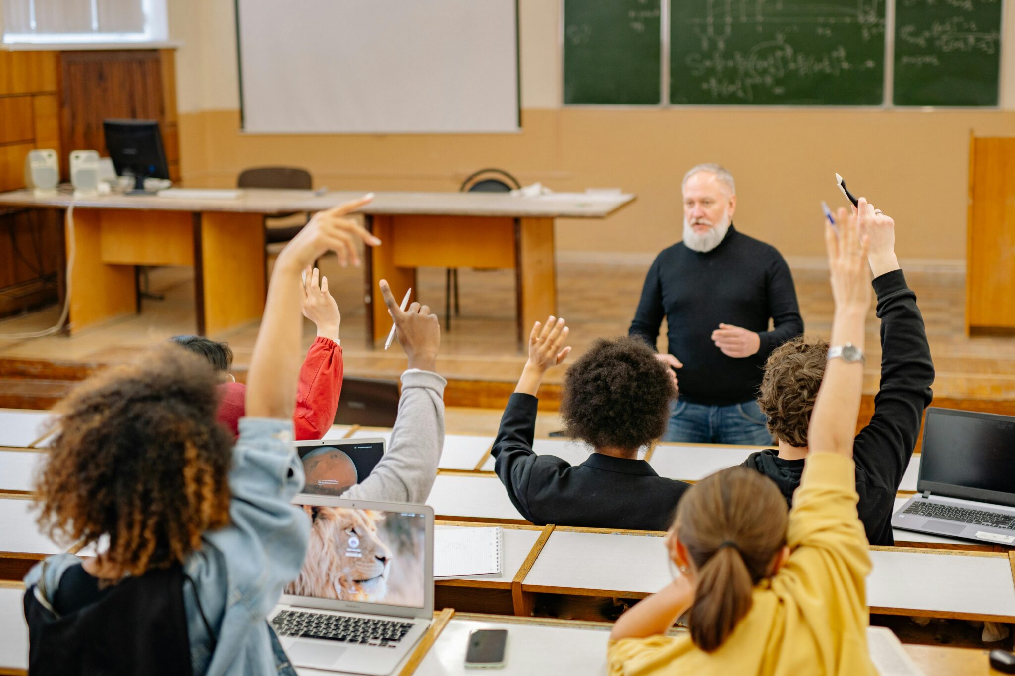A photo of classroom where the teacher is asking a question, and the students are raising their hands to respond.