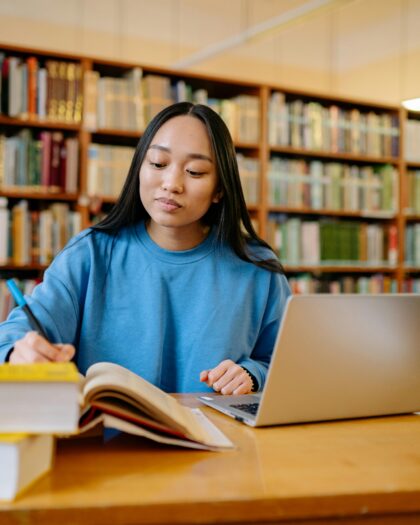 A young woman studying in a library, sitting at a table with an open laptop and notebook, writing notes while surrounded by books.
