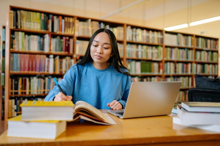 A young woman studying in a library, sitting at a table with an open laptop and notebook, writing notes while surrounded by books.