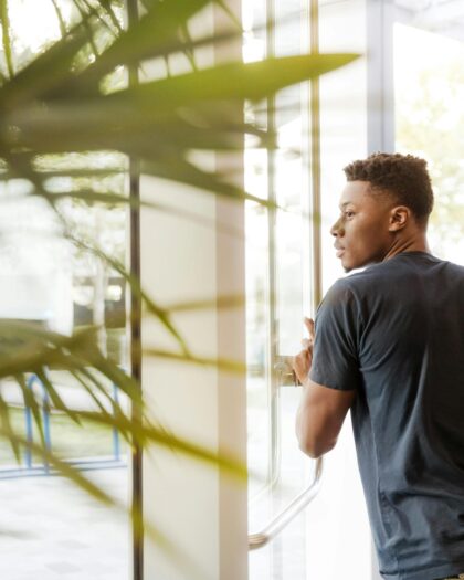 Man Looking Outside Window Carrying Black and Brown Backpack While Holding His Hand on Window