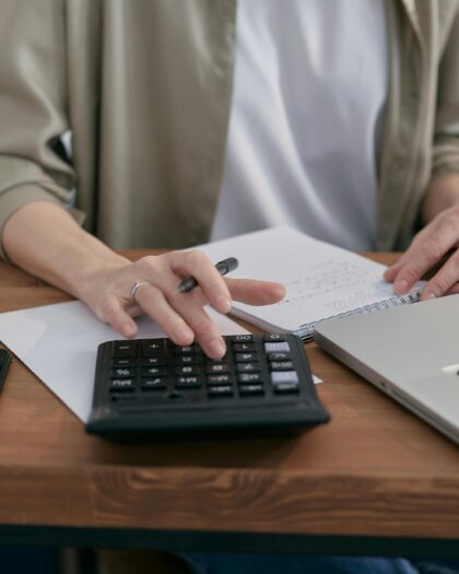 A Person Sitting a Wooden Table with a Notepad and Laptop Using a Calculator