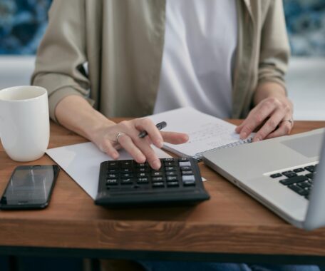 A Person Sitting a Wooden Table with a Notepad and Laptop Using a Calculator