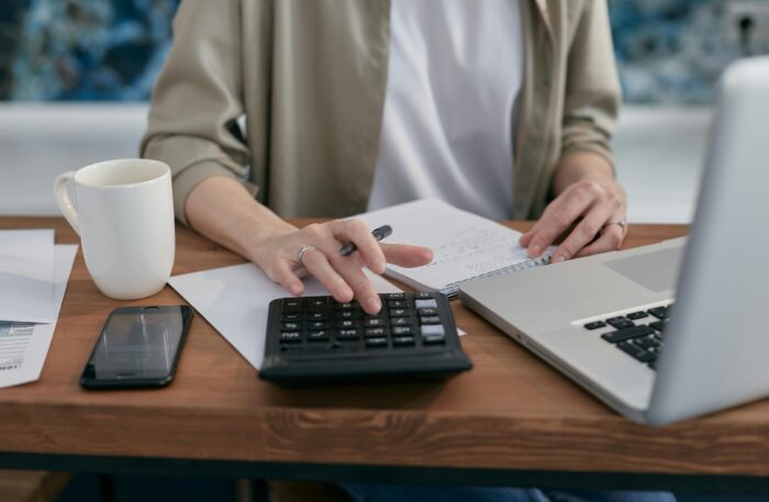 A Person Sitting a Wooden Table with a Notepad and Laptop Using a Calculator