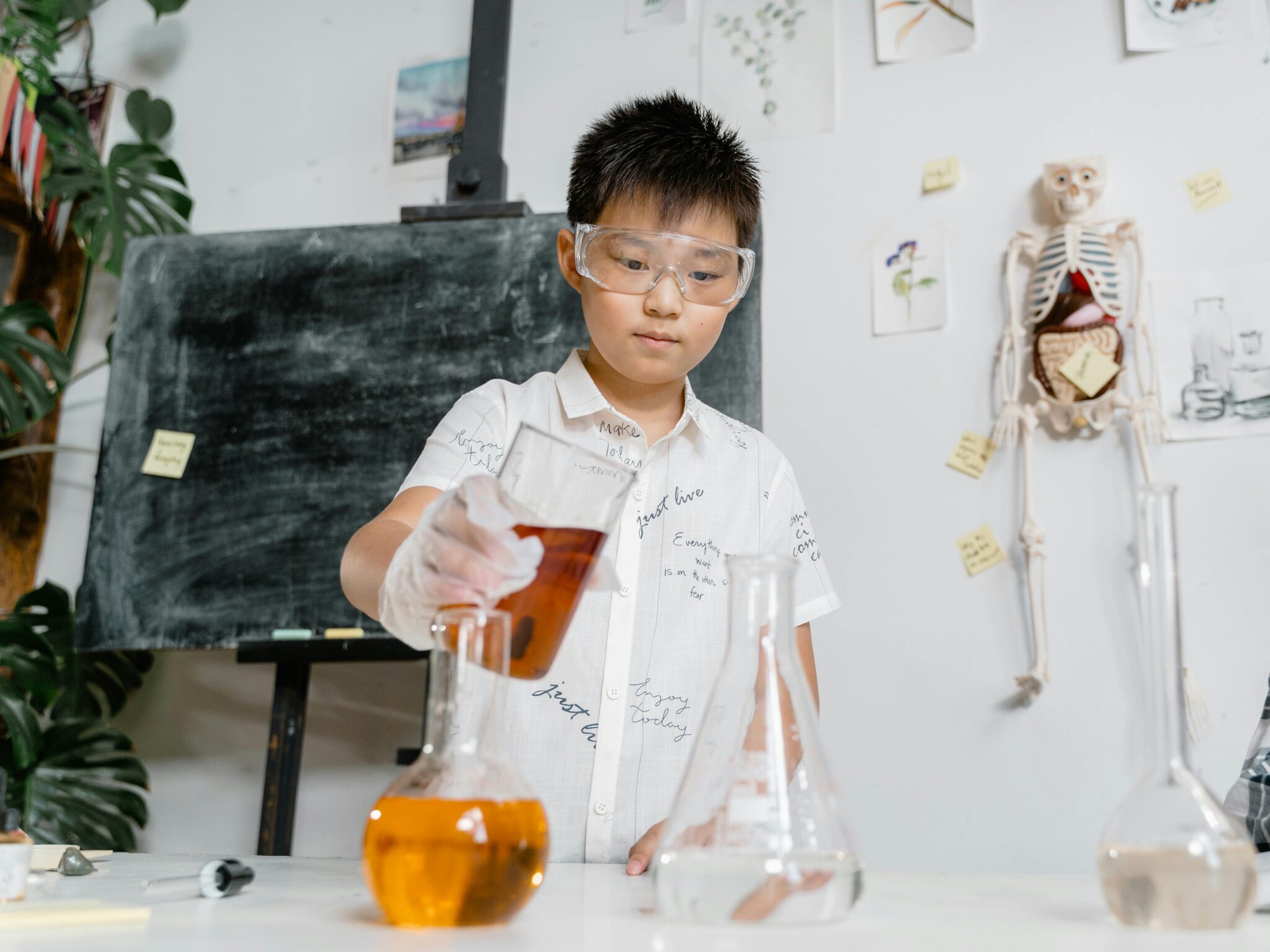 A child in goggles and a lab coat pours amber liquid into a beaker in a science classroom. A blackboard and skeleton model are in the background.